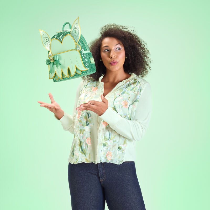 Woman wearing a green top and blue jeans, throwing the Loungefly Tinker Bell holiday mini backpack up in the air. She stands against a green background.