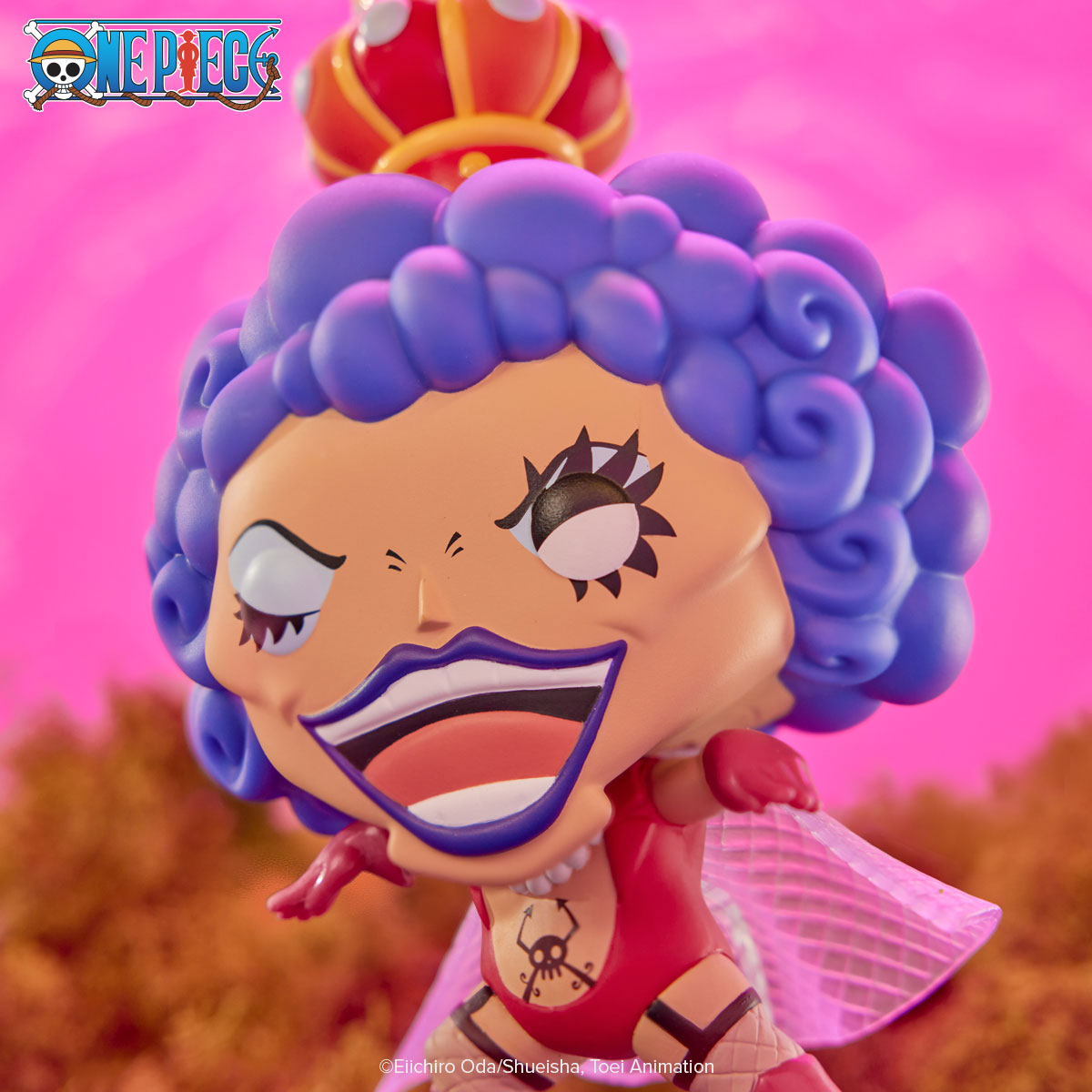 This lifestyle photo shows POP! Emporio Ivankov in action, with a winking eye. POP! Emporio Ivankov wears signature fishnet stockings and red body suit, displaying tattoos. A pink cape "flutters" behind. The sky behind this figure is cotton-candy pink and vague images of vegetation are in the background.