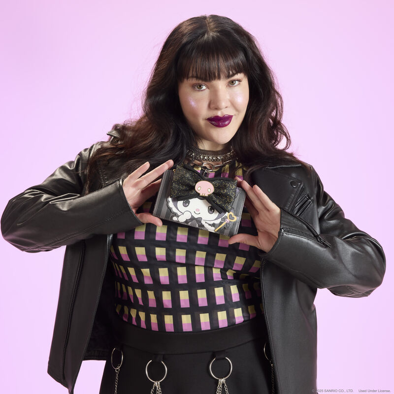 Woman standing against a purple-pink background, wearing a black, pink, and yellow top and black leather jacket, holding the Loungefly Kuromi 20th anniversary flap wallet. 