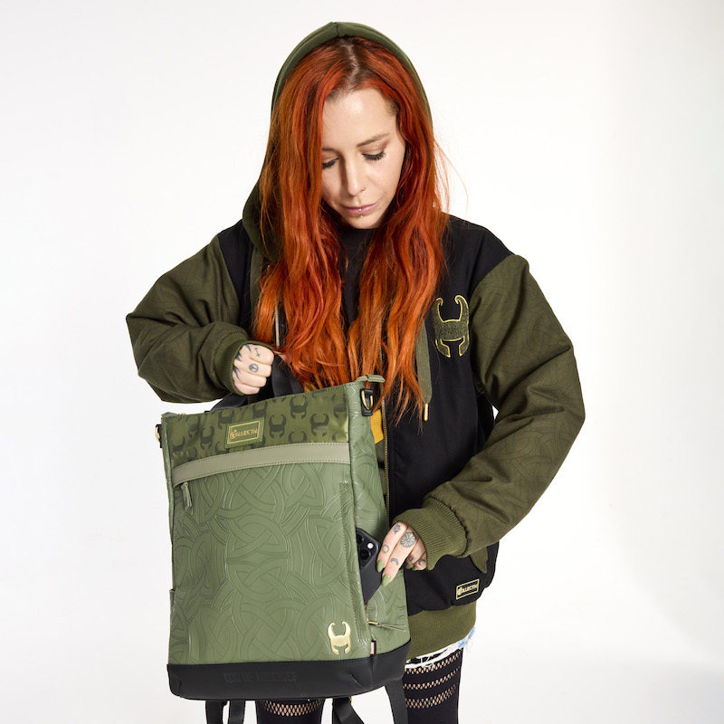 Red-headed woman wearing the Loungefly COLLECTIV Marvel Loki The WEEKENDR Hooded Jacket and holding the Loungefly COLLECTIV Marvel Loki The CREATIV Convertible Tote Bag with one hand and sliding a phone into a hidden pocket on the side with the other hand