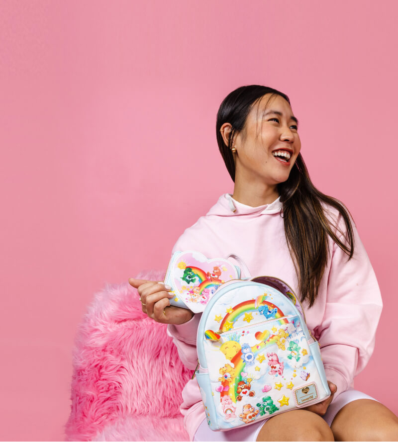 Pink colored background with model holding Loungefly Care Bears Bags and Wallets. Click to shop now!