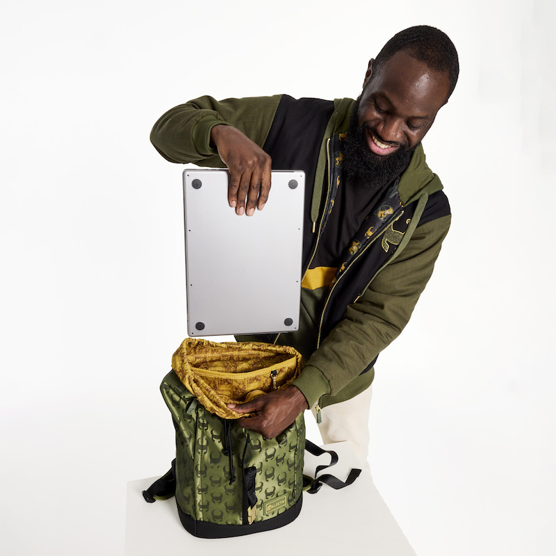 Man wearing the Loungefly COLLECTIV Marvel Loki The WEEKENDR Hooded Jacket and the ORIGINL tee while holding the TRAVELR full-size backpack open and putting a silver laptop into it.