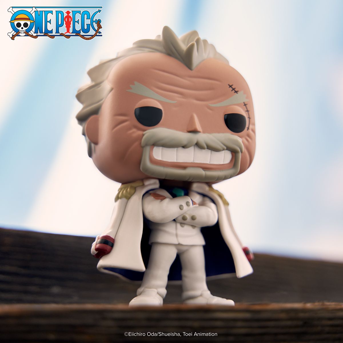 This photo displays an image of exclusive POP! Monkey D. Garp, dressed in his white suit, with his arms crossed in front. He's standing on a high platform of sorts with wood grain/shingles. In the background, there's an impression of a blue and white "sky" background.